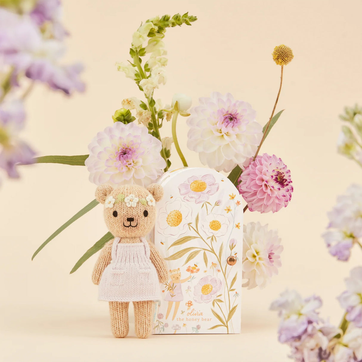 Knitted bear toy with a pale pink dress surrounded by flowers on a beige background