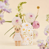Knitted bear toy with a pale pink dress surrounded by flowers on a beige background