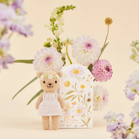 Knitted bear toy with a pale pink dress surrounded by flowers on a beige background