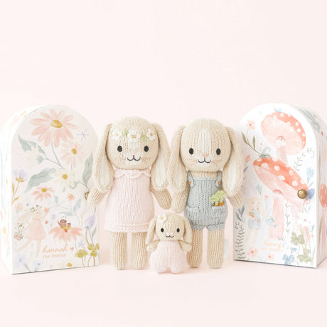 Three knitted bunny dolls one in a pink dress with a flower chain on her head. Next to her bunny toy in a blue dunagree outfit with carrots in his pocket and a baby bunny in between them in pink. They stand in front of watercoloured gift boxes