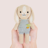 nitted bunny toy in a blue dunagree outfit with carrots in his pocket held by hands against a light pink background