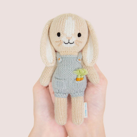 nitted bunny toy in a blue dunagree outfit with carrots in his pocket held by hands against a light pink background