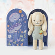 Knitted bunny toy in a blue dunagree outfit with carrots in his pocket with a decorative round arch shaped box featuring floral and mushroom design on a light background.