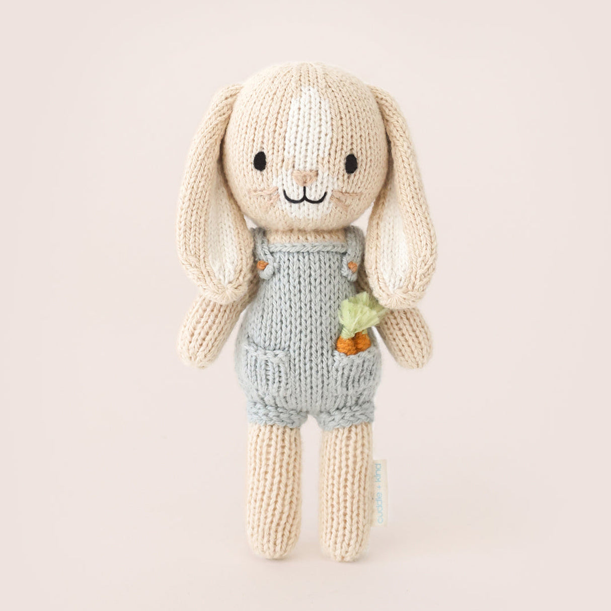 knitted bunny toy in a blue dunagree outfit with carrots in his pocket on a light brown background