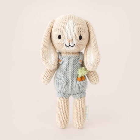 knitted bunny toy in a blue dunagree outfit with carrots in his pocket on a light brown background