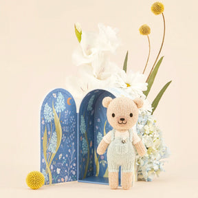 Knitted teddy bear in overalls standing next to gift bow with floral illustrations.