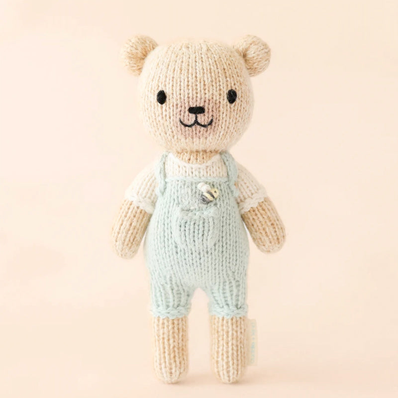 Knitted teddy bear wearing light blue overalls on a beige background with knitted bumble bee