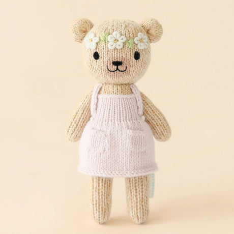Knitted teddy bear wearing a pink dress with a flower headband on a beige background