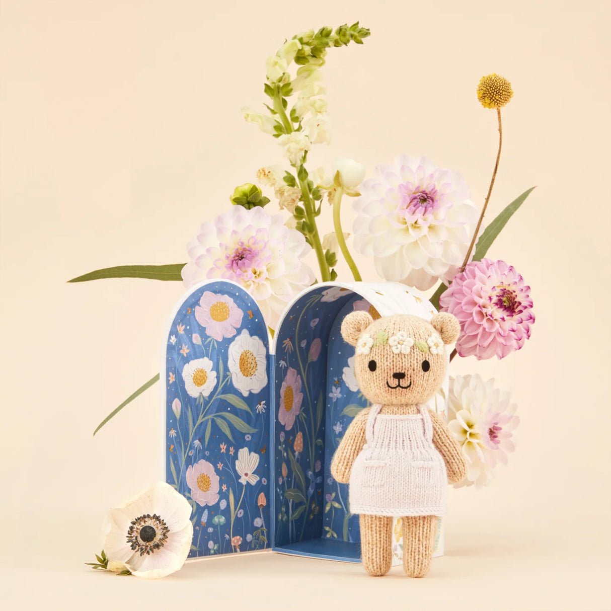 Small teddy bear in a light pink dress standing next to floral arched box with flowers on a beige background