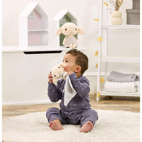 Baby sitting holding a Harry Potter themed Dobby comforter, featuring a plush Dobby the house-elf on a grey velvety blanket, with a neutral color palette. 