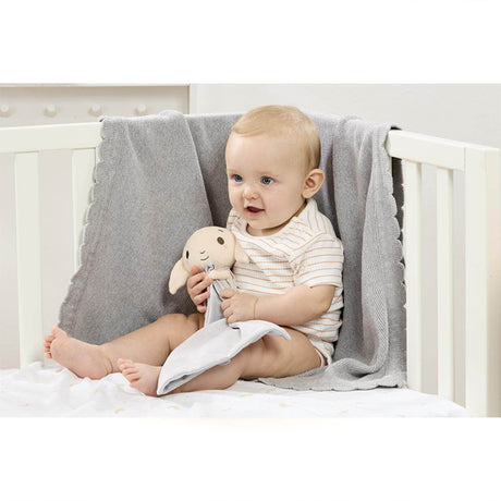 Baby sitting in a crib holding a Harry Potter themed Dobby comforter, featuring a plush Dobby the house-elf on a grey velvety blanket.