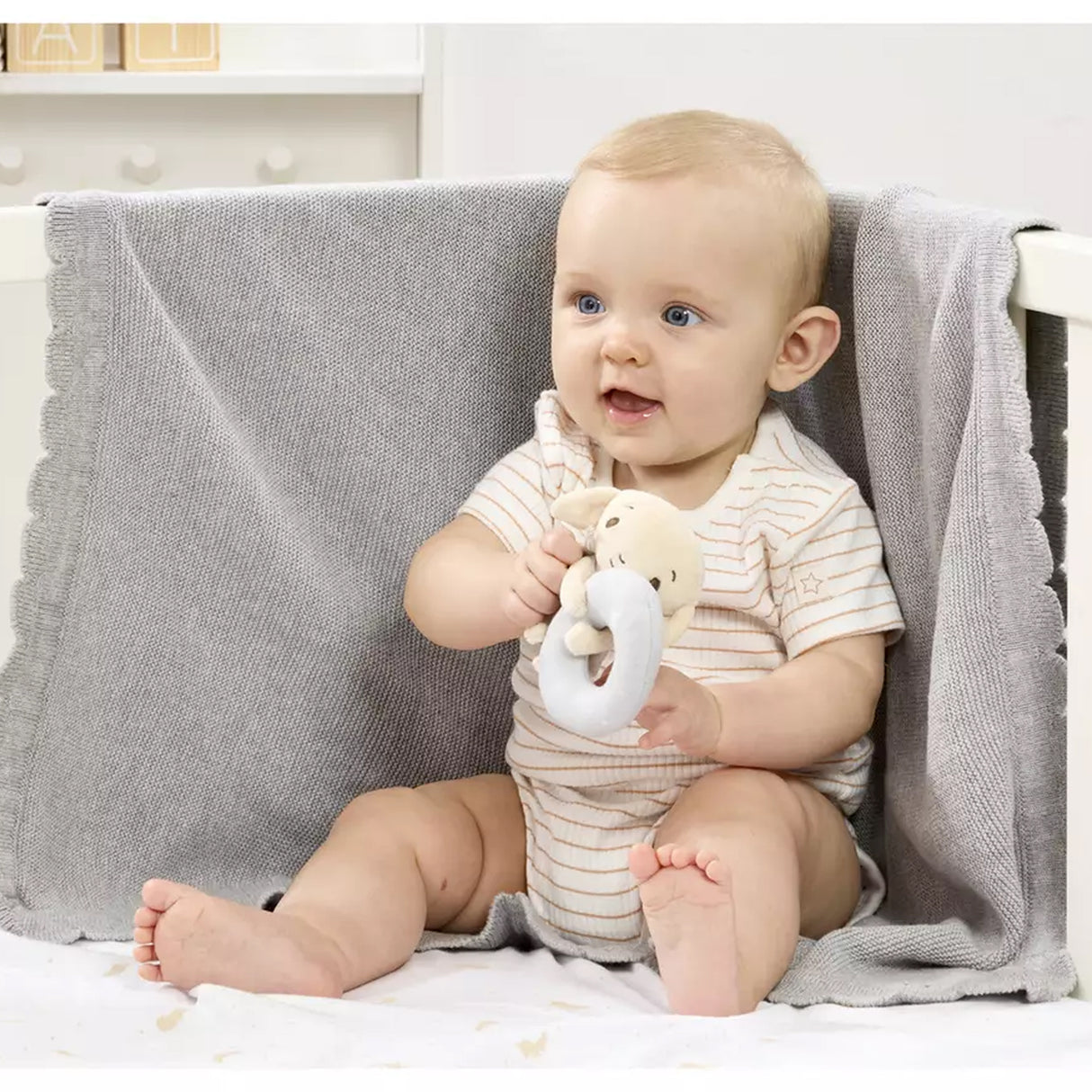 Baby sitting on a bed holding a Plush Dobby ring rattle toy, with a neutral background