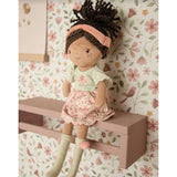 Soft doll with floral pink and sage green dress, pink headband and curly brown hair tied in a ponytail against a floral bird wallpapered background