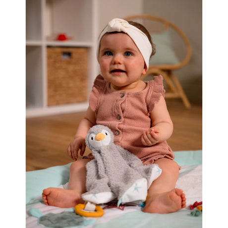 A baby sitting on a blanket holding a grey soft penguin soother.
