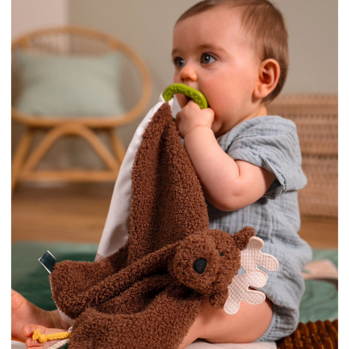 A baby sitting on the floor holding a brown plush moose security blanket with a green teething ring, in a softly lit nursery setting.