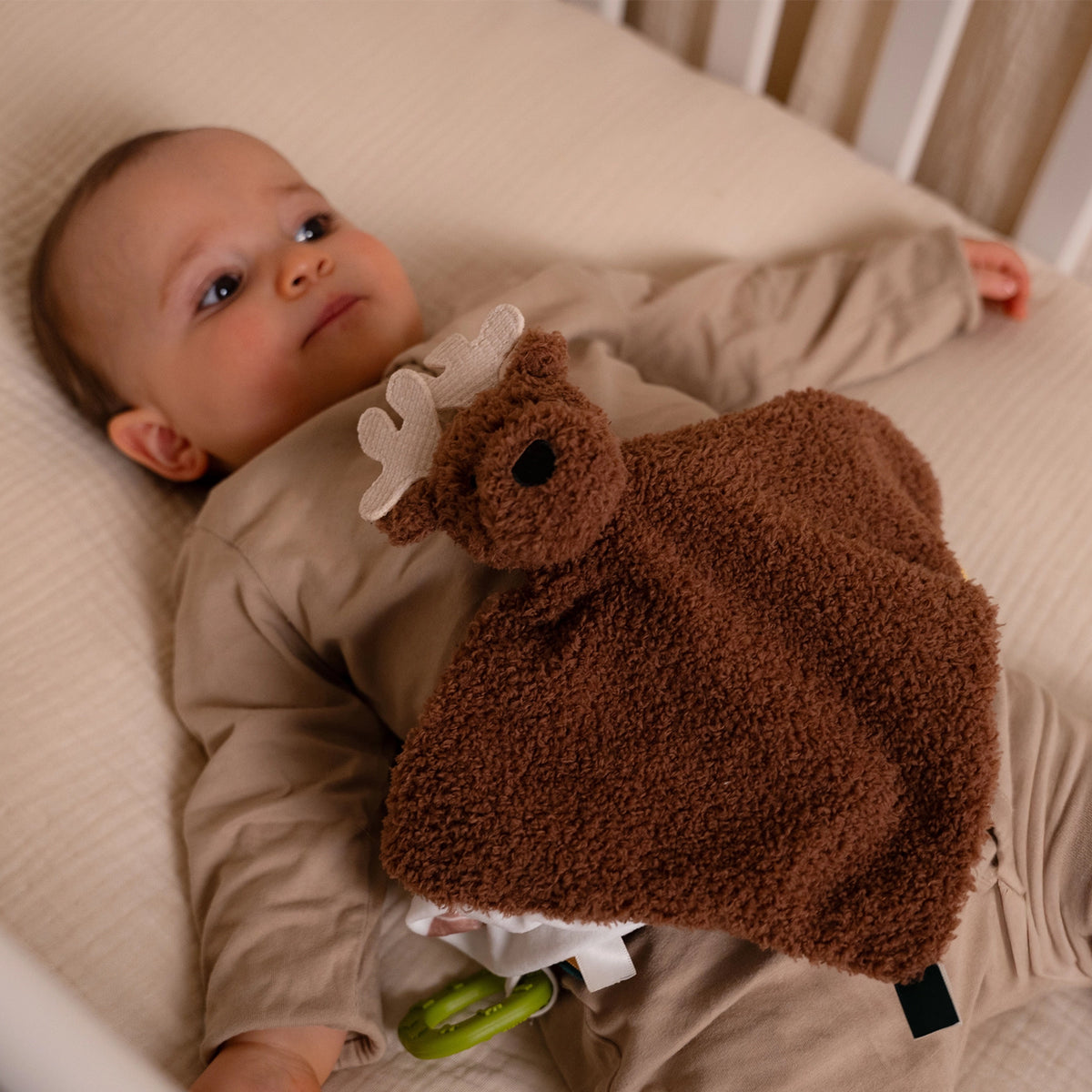 A baby lying on a cream-coloured bed, holding a brown fluffy security blanket shaped like a moose with a white antler and a green teething ring attached.