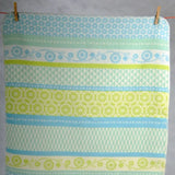 A cotton blanket with retro floral and geometric pattern in light blue and lime green, hanging from two wooden pegs.