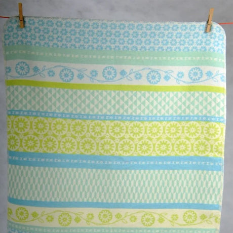 A cotton blanket with retro floral and geometric pattern in light blue and lime green, hanging from two wooden pegs.