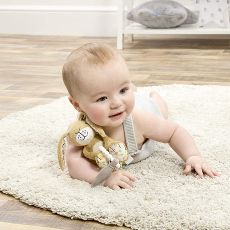Baby sitting on a rug with a Plush nutbrown hare rabbit toy holding a leaf and silicon teether ring on a white background