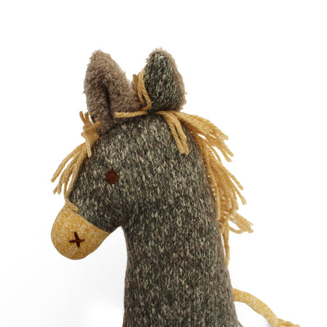 A close-up of a woolly horse knitted soft toy, showing its textured gray and beige body, yellow yarn mane, and embroidered details on a plain white background.