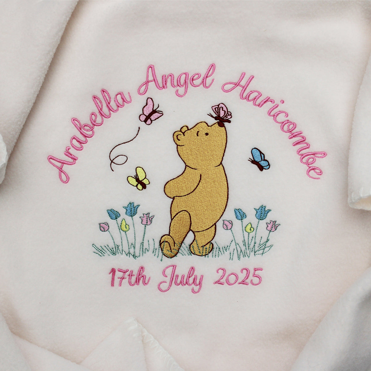 Close-up of a cream-coloured baby blanket with a personalised design in the centre, featuring Winnie the Pooh, pink name and date of birth embroidered and small blue and yellow flowers.