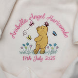 Close-up of a cream-coloured baby blanket with a personalised design in the centre, featuring Winnie the Pooh, pink name and date of birth embroidered and small blue and yellow flowers.