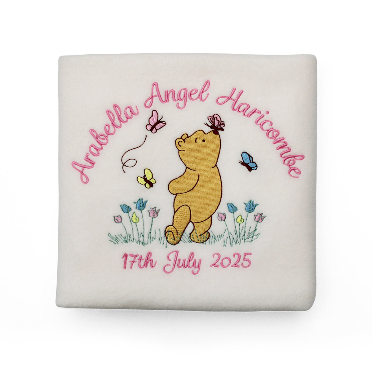 A cream-coloured baby blanket with an embroidered personalised design in the centre, featuring Winnie the Pooh with a butterfly on his nose, pink name and date of birth and small blue and yellow flowers.