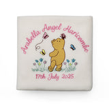 A cream-coloured baby blanket with an embroidered personalised design in the centre, featuring Winnie the Pooh with a butterfly on his nose, pink name and date of birth and small blue and yellow flowers.
