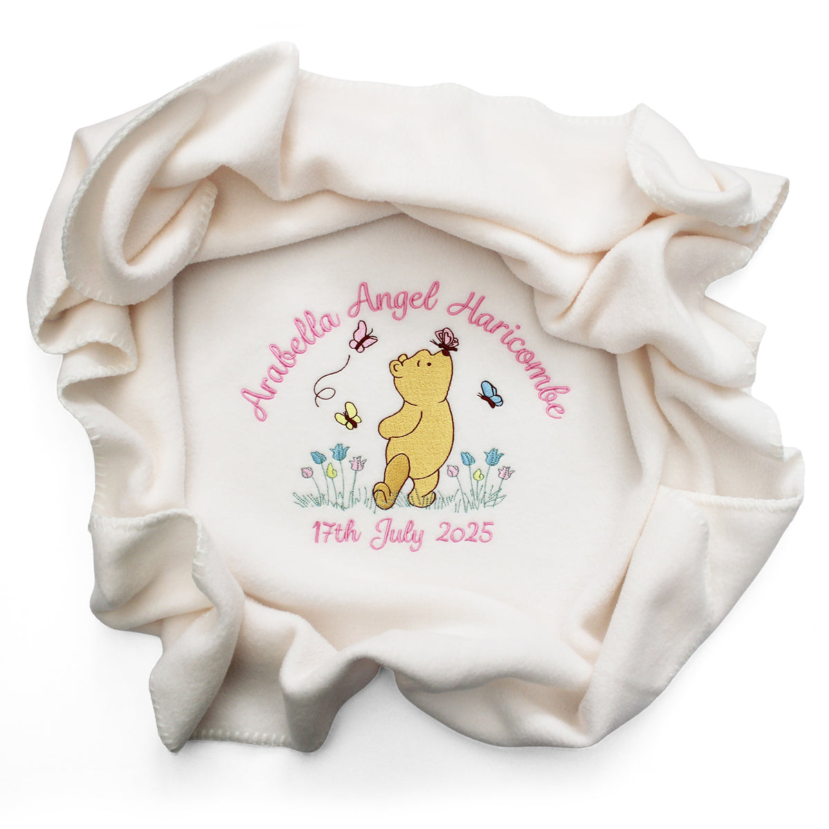A cream-coloured baby blanket with a personalised design in the centre, featuring Winnie the Pooh, pink name and date of birth embroidered and small blue and yellow flowers.