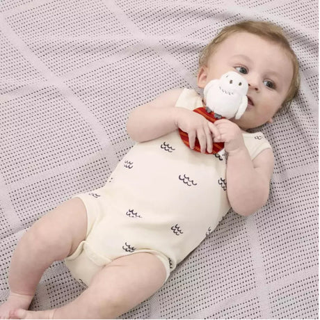 Baby lying on a textured blanket holding a A plush Harry Potter Hedwig baby ring rattle rattle with a white owl on top sitting on a red and orange striped ring.