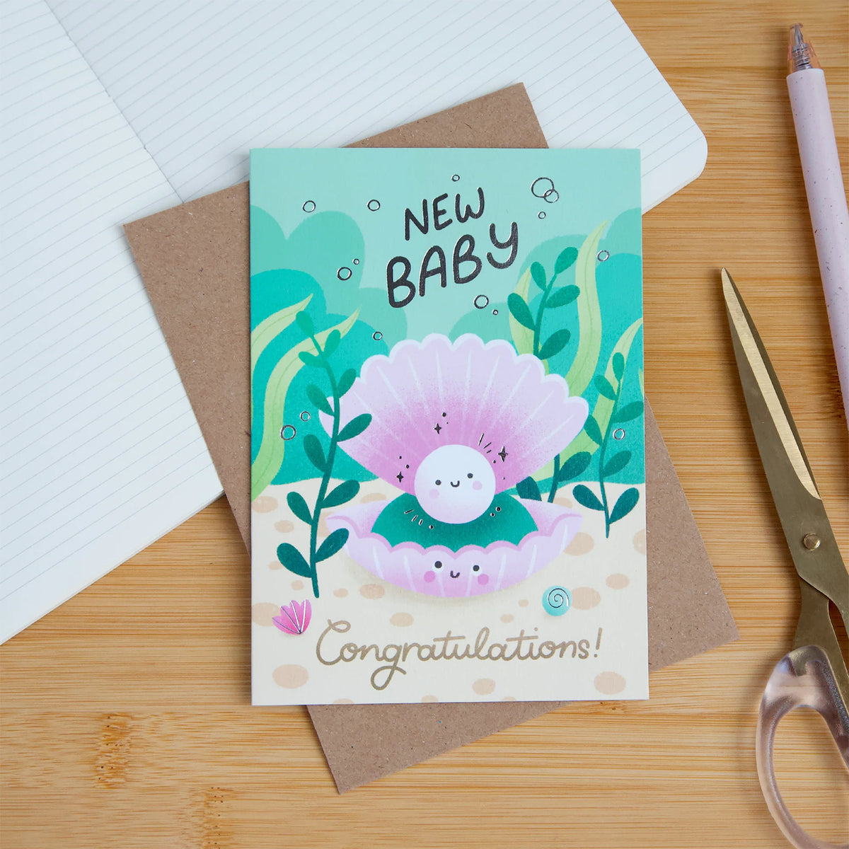A 'New Baby' congratulations card featuring a cute cartoon baby in a pink clam shell, displayed on a wooden desk with a brown envelope, notebook, scissors, and a pen.