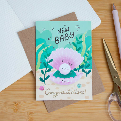 A 'New Baby' congratulations card featuring a cute cartoon baby in a pink clam shell, displayed on a wooden desk with a brown envelope, notebook, scissors, and a pen.