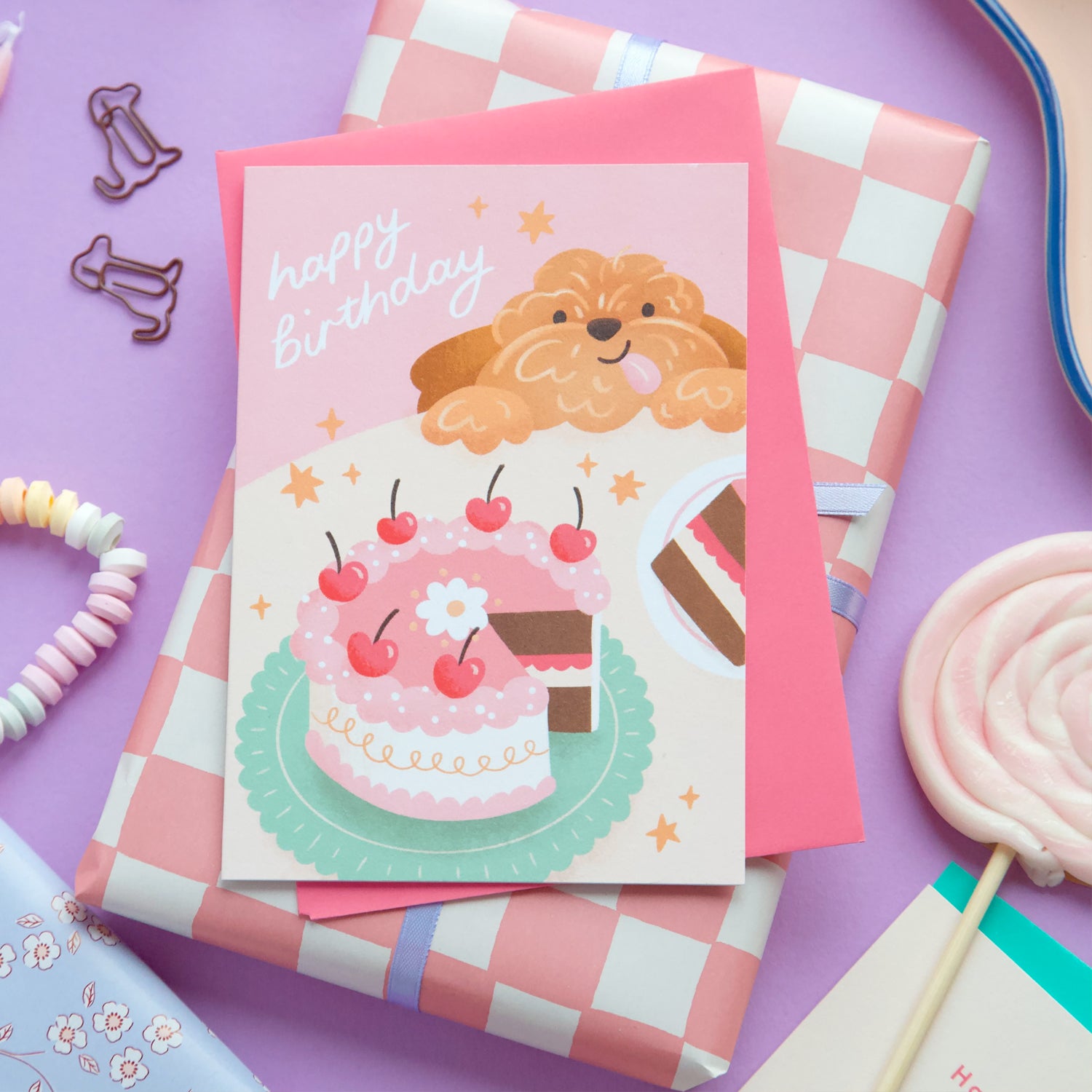 Birthday card with a dog on top of a cake illustration, surrounded by party supplies on a purple background.