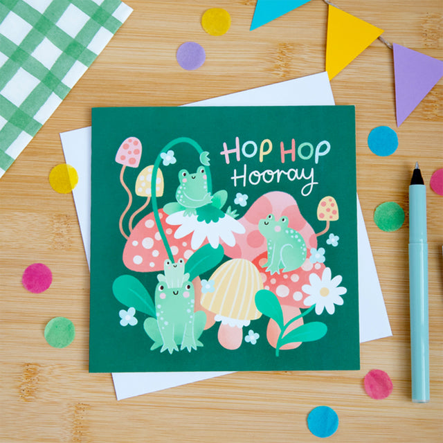 Colorful greeting card with frogs and text 'Hop Hop Hooray' on a wooden surface with confetti and a pen.