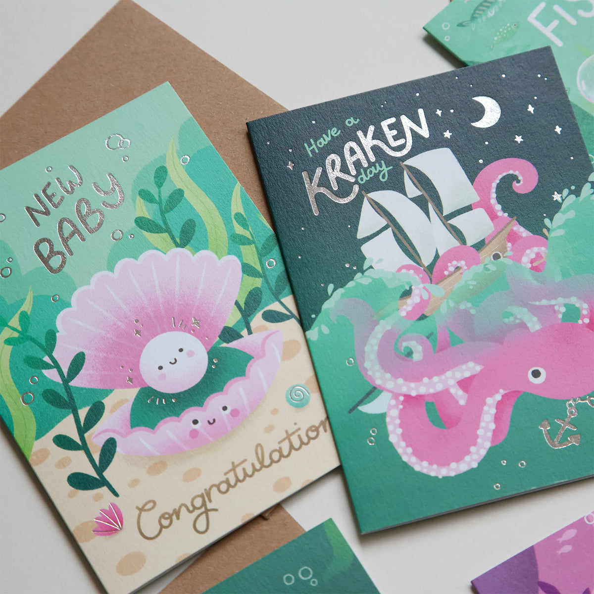 Two illustrated greeting cards arranged on a light surface with brown envelopes. One card says "NEW BABY" with a pink clam character and "Congratulations" text; the other says "Have a KRAKEN day" with a pink octopus and white ship under a crescent moon.