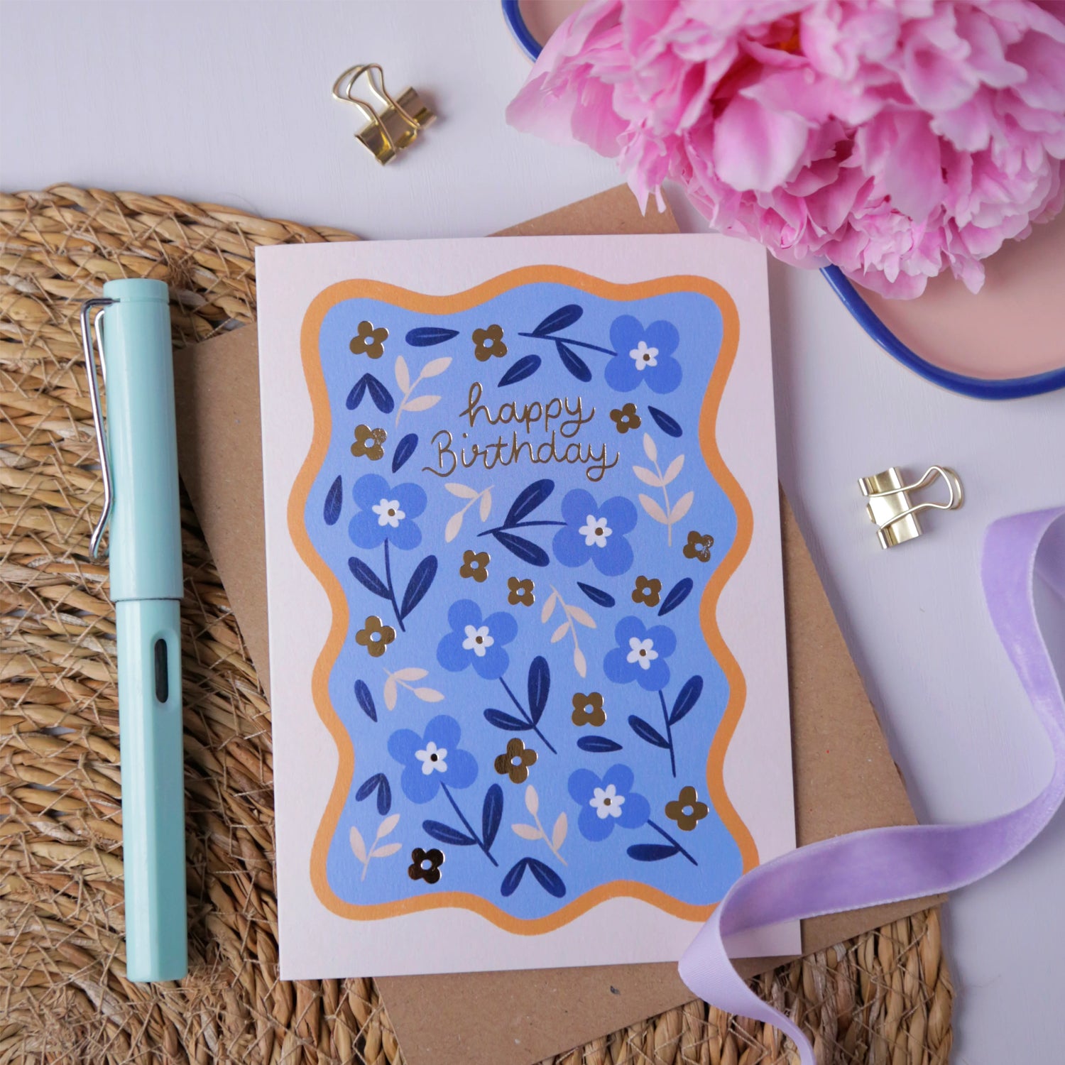 Hopscotch Paper Co. blue floral Happy Birthday card with gold foil text, styled with pen and pink flowers on rattan mat