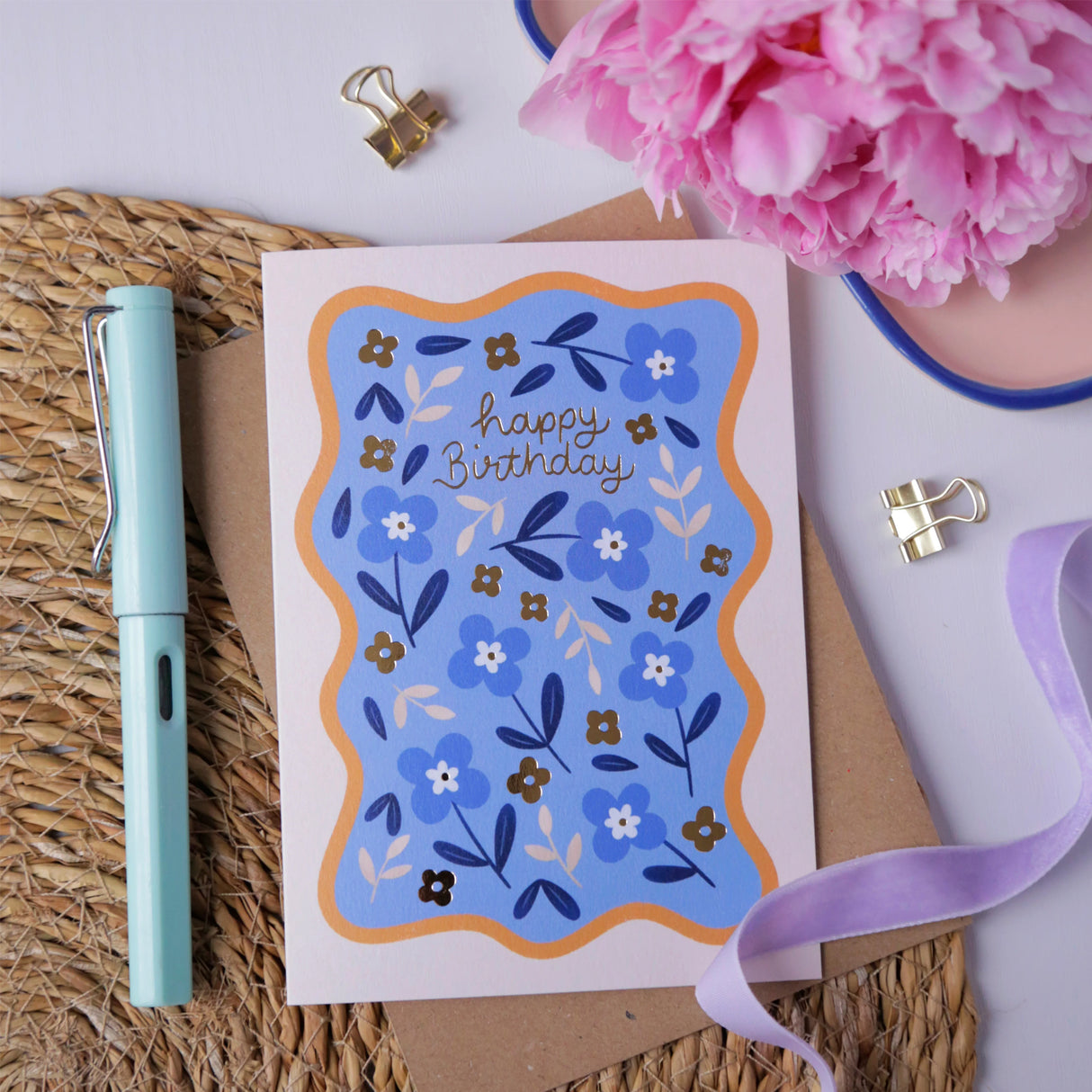 Hopscotch Paper Co. blue floral Happy Birthday card with gold foil text, styled with pen and pink flowers on rattan mat