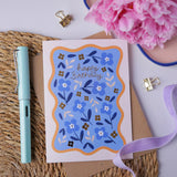 Hopscotch Paper Co. blue floral Happy Birthday card with gold foil text, styled with pen and pink flowers on rattan mat