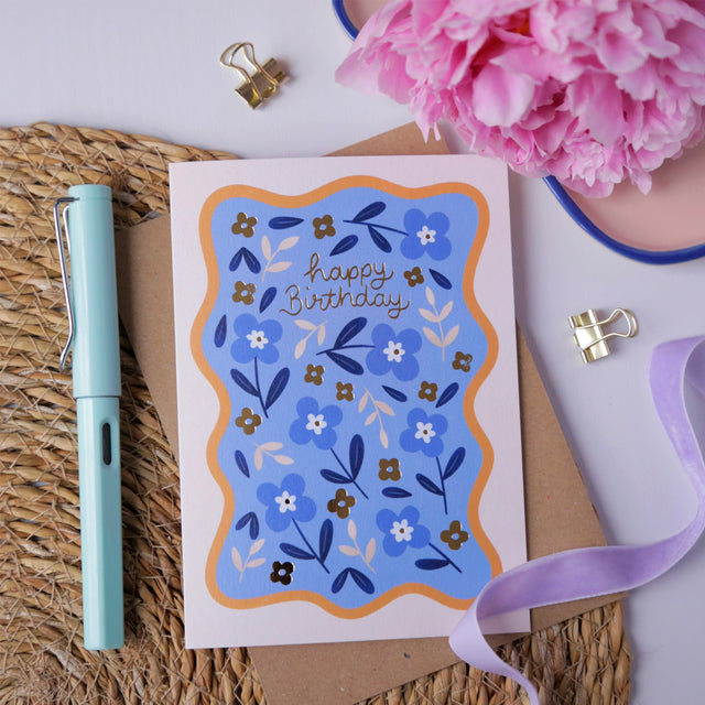 Hopscotch Paper Co. blue floral Happy Birthday card with gold foil text, styled with pen and pink flowers on rattan mat