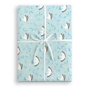 A Patterned blue gift wrap present with white storks holding a blue bundle and blue leaves with purple flowers wrapped with white satin ribbon