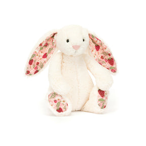 A small plush toy resembling a cream-colored bunny with soft cotton ears and feet, featuring a strawberry and cream flower pattern on a pink backdrop.