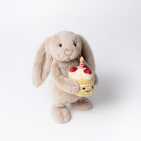 A beige plush toy rabbit standing holding a birthday cake with a single candle on top.