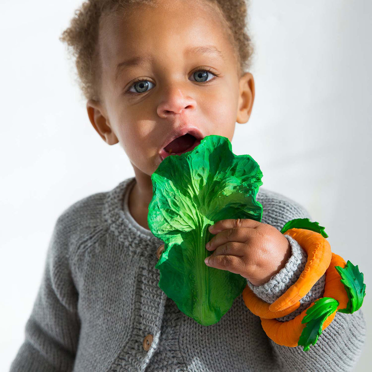 Child holding a green leafy kale rubber teether toy with an orange and green toy on wrist against a white background