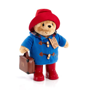 Plush toy Paddington Bear with red hat, blue coat with wooden toggles and P.B embroidered in yellow, red boots, and holding brown suitcase on white background