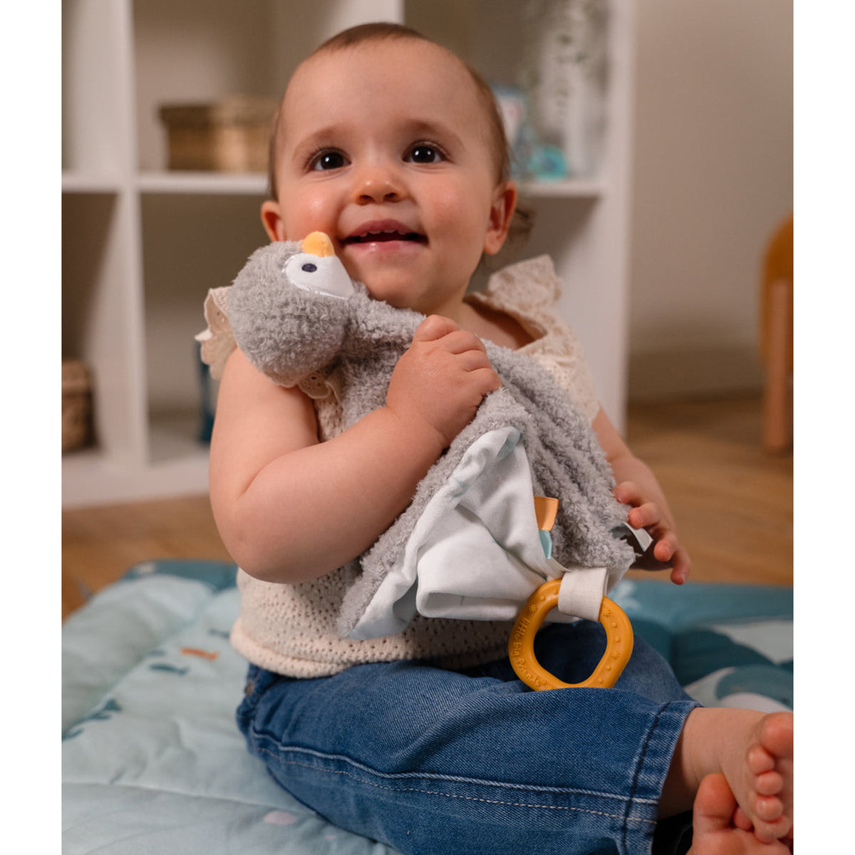 A baby sitting holds a grey plush penguin toy with a white fabric belly and a yellow ring toy attached