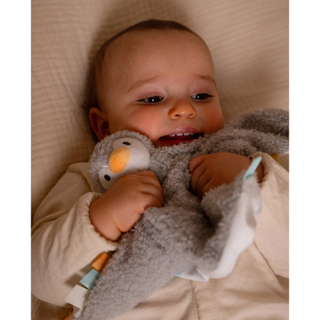 A baby lying holding a gray plush penguin toy with a white belly, orange beak
