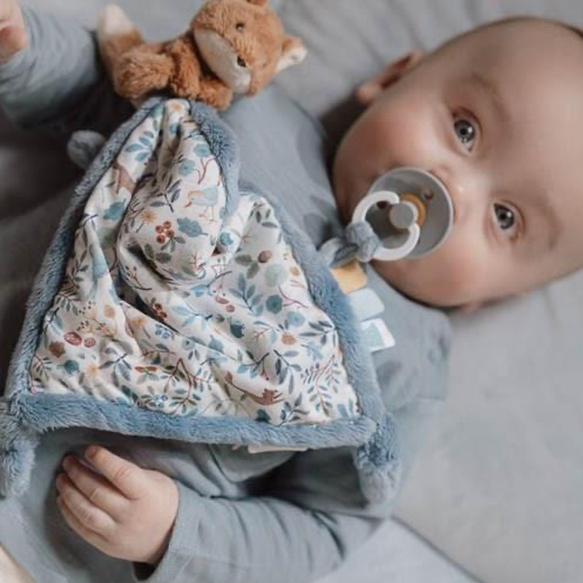 Baby lying on a bed with a woodland print soother blanket with fox toy, and pacifier.