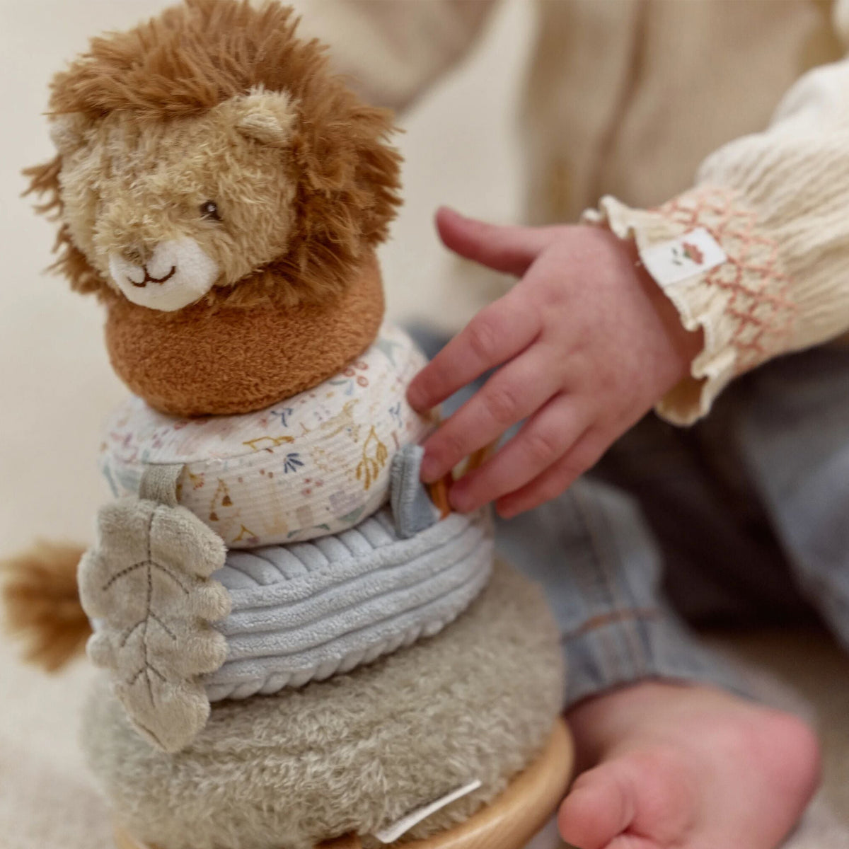 A close-up of a baby playing with a plush toy stack on a wooden base. The toy features a brown lion head on a brown plush base, stacked with a patterned fabric cushion, a light blue ribbed cushion, and a gray textured cushion.