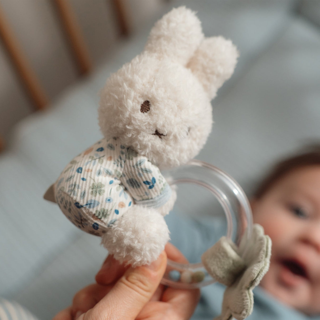 White fluffy miffy toy with a floral leaf pattern held by a hand, with a blurred baby in the background.