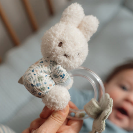 White fluffy miffy toy with a floral leaf pattern held by a hand, with a blurred baby in the background.